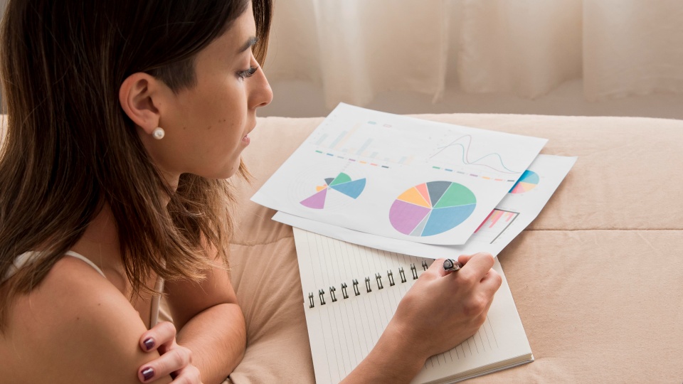 Uma mulher está sentada à mesa, concentrando-se em escrever em um caderno. À sua frente estão papéis com gráficos coloridos relacionados ao programa acelerar a economia. Ela está vestindo um top sem mangas e brincos de pérolas. A luz natural ilumina a cena através de uma cortina transparente ao fundo. - Efacont