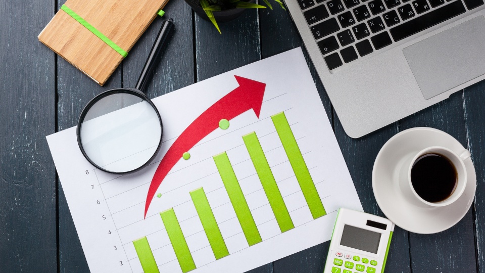 Um gráfico de barras com barras verdes e uma seta vermelha para cima é colocado sobre uma mesa de madeira escura. Os itens ao redor do gráfico incluem uma lupa, um caderno de madeira, um vaso de planta, um laptop, uma xícara de café e uma calculadora verde – sugerindo o cenário para analisar estratégias para programar acelerar a economia. - Efacont