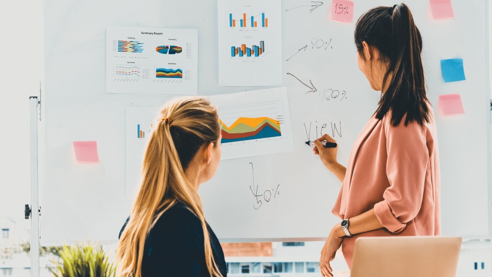 Duas mulheres colaboram em frente a um quadro branco adornado com tabelas coloridas, gráficos e post-its. Um fica de pé, segurando um marcador, fazendo anotações no quadro branco. O outro fica sentado de frente para o quadro, participando ativamente. O cenário parece ser um ambiente de escritório focado em consultoria estratégica. - Efacont