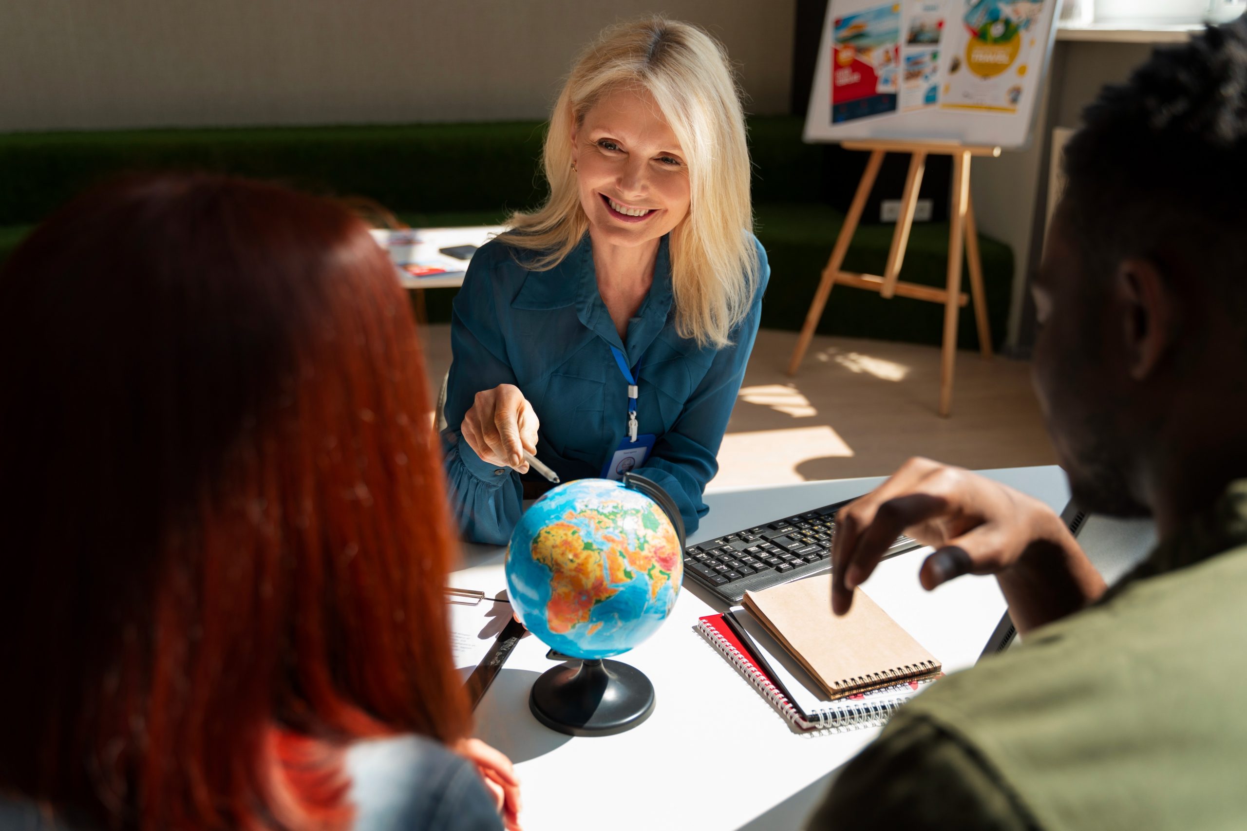 Uma mulher loira discute planos de viagem com duas pessoas em uma consultoria em turismo. Ela está sorrindo e apontando para um globo sobre a mesa, cercado por cadernos, uma calculadora e folhetos de viagens. O cenário parece ser um escritório ou agência de consultoria em turismo. - Efacont