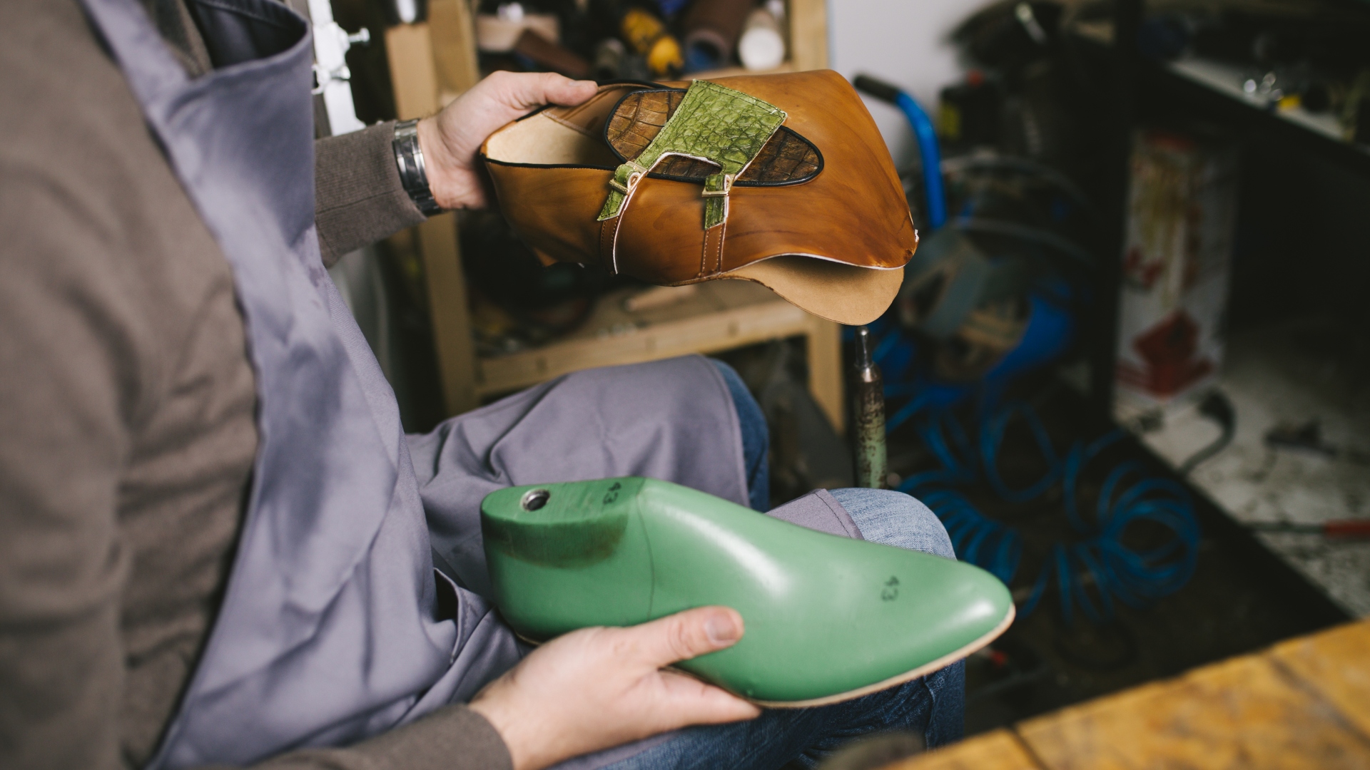 Um sapateiro está sentado, segurando um sapato feito de couro marrom e verde em uma das mãos e um sapato de plástico verde na outra. O artesão, de avental cinza, está rodeado de diversas ferramentas e equipamentos em um ambiente de oficina que valoriza o desenvolvimento sustentável. - Efacont