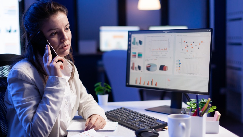 Uma mulher está sentada em uma mesa, segurando um telefone no ouvido com a mão esquerda enquanto olha para um monitor de computador que exibe vários gráficos relacionados ao SAF-T. Ela tem um bloco de notas à sua frente e um teclado, um mouse, um pequeno vaso de planta e uma caneca sobre a mesa. O fundo está desfocado, com uma lâmpada visível. - Efacont