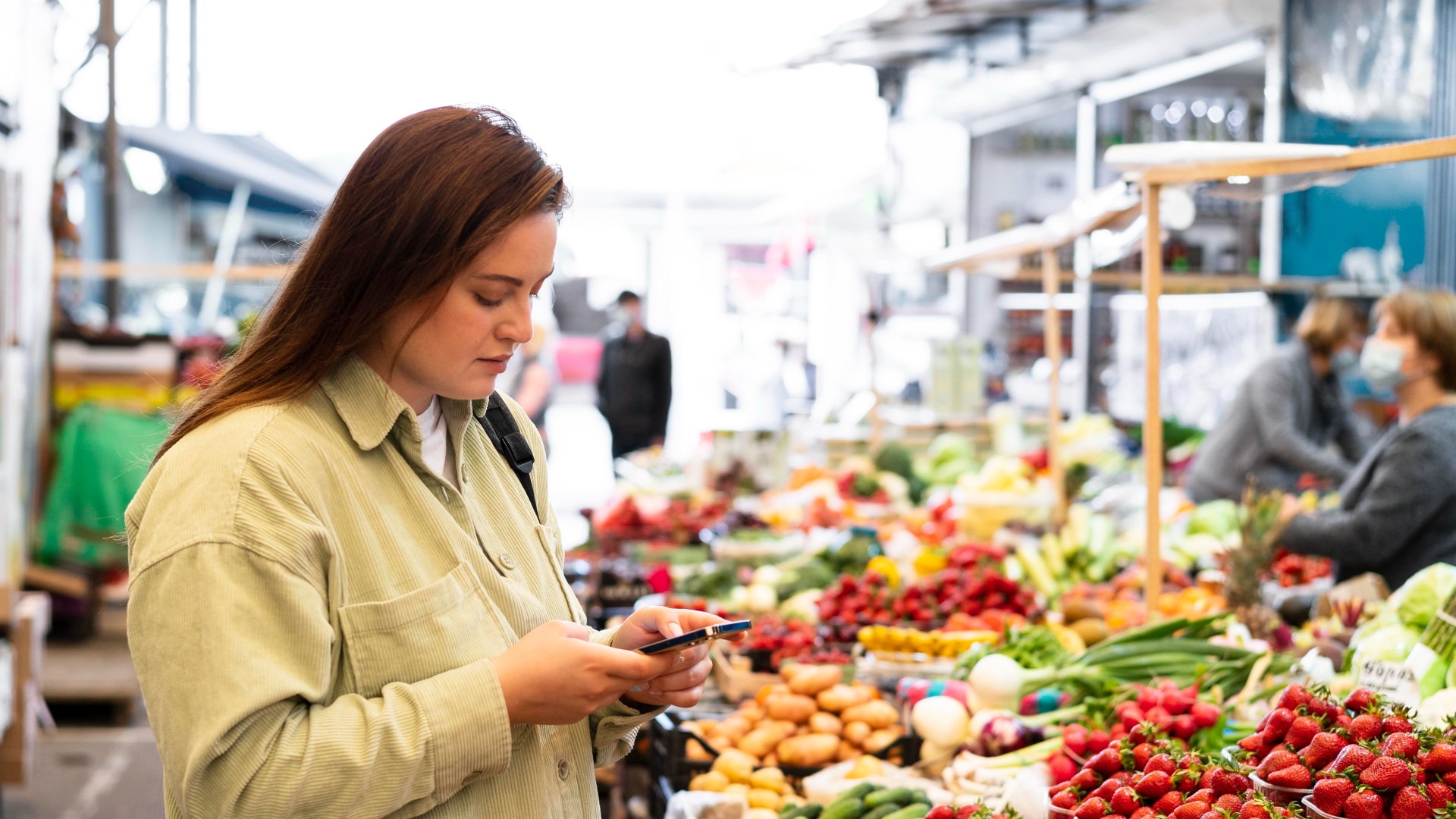 Uma mulher com longos cabelos castanhos, vestindo uma camisa verde clara, está em um movimentado mercado ao ar livre repleto de frutas e vegetais coloridos. No meio do vibrante Setor Alimentar Português, ela olha para o seu smartphone. Outros frequentadores do mercado e vendedores são visíveis ao fundo, comprando e organizando produtos. - Efacont