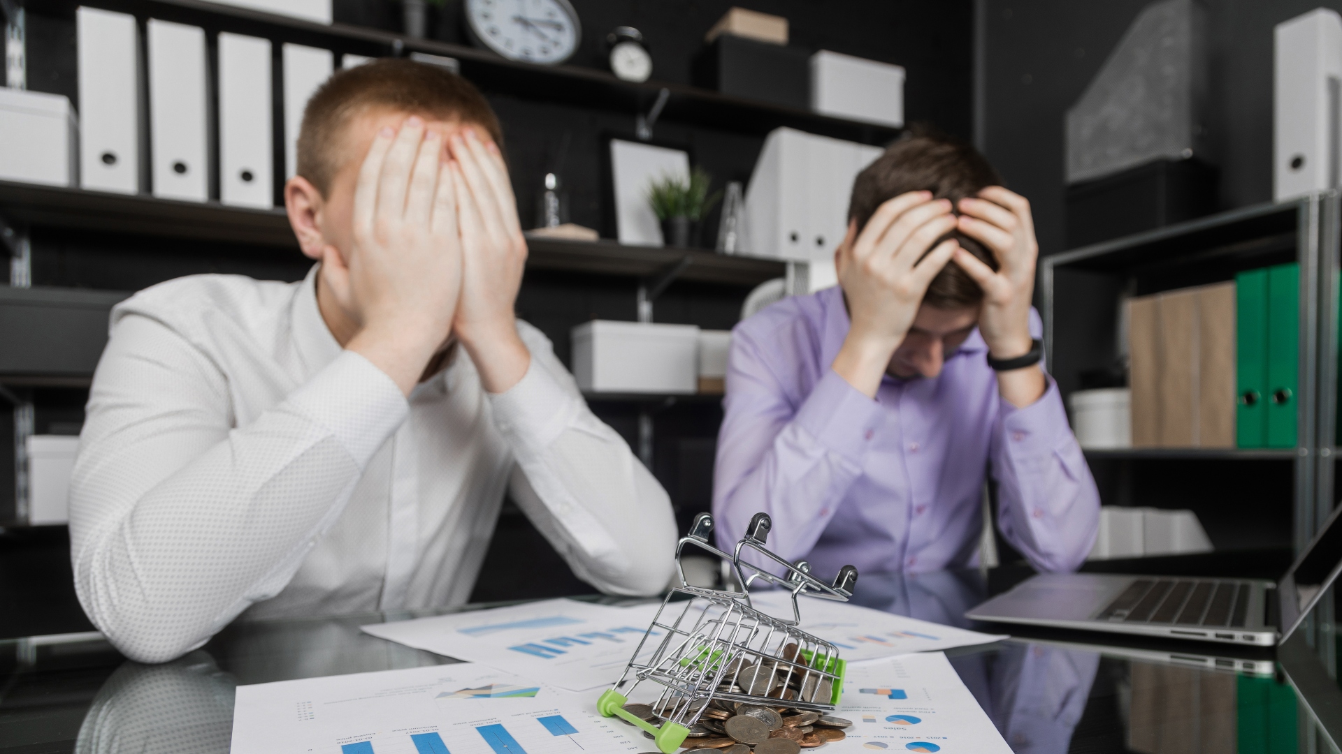 Dois homens em um escritório sentam-se à mesa com as cabeças entre as mãos, personificando as tensões da dependência financeira. Um pequeno carrinho de compras cheio de moedas está virado sobre documentos com gráficos. Prateleiras com fichários e um relógio aparecem ao fundo, enquanto um laptop aberto está diante de um homem. - Efacont