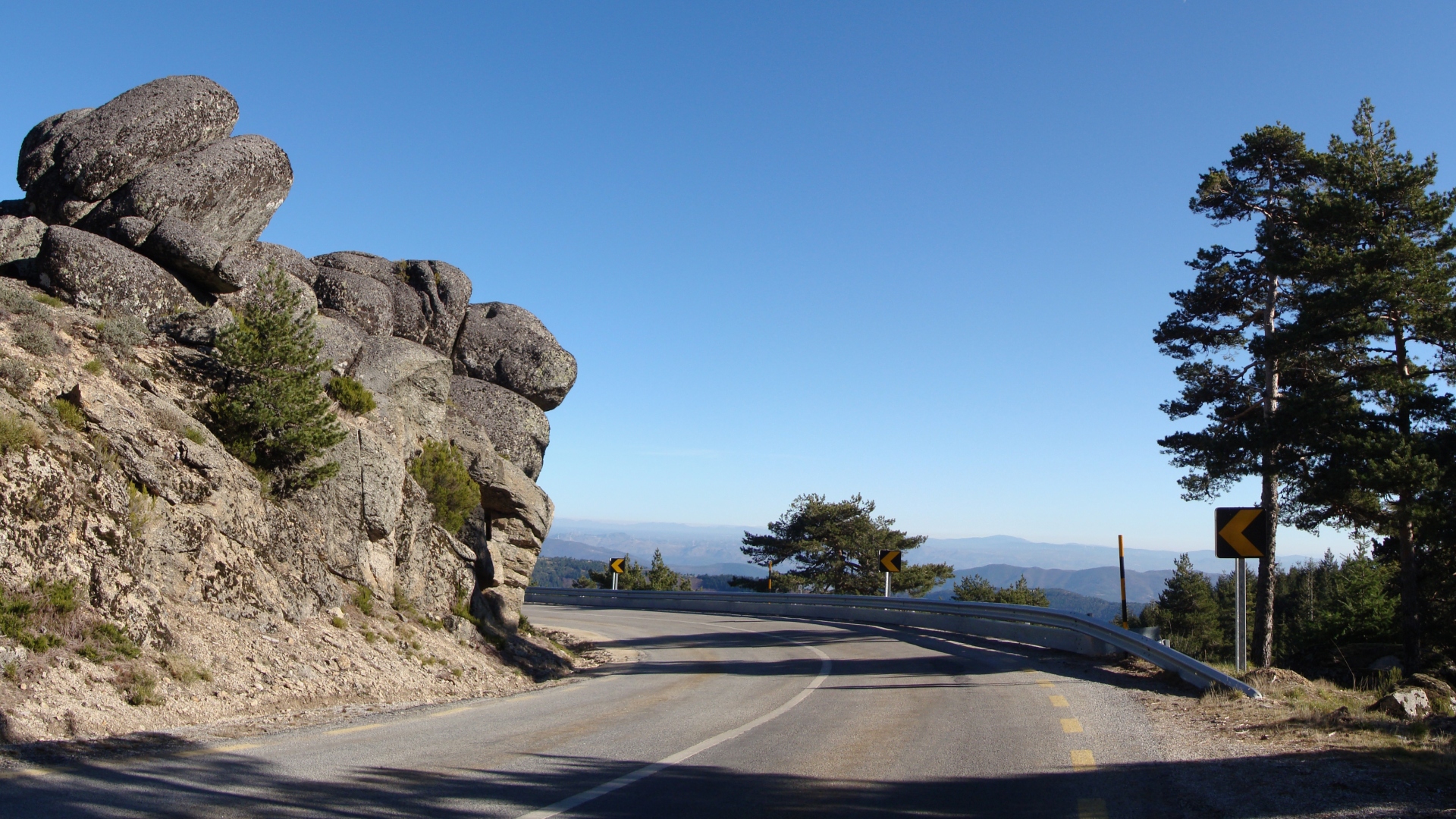 Uma estrada de montanha cênica serpenteia em uma curva, passando por grandes formações rochosas escarpadas à esquerda. Guarda-corpos delimitam a estrada para segurança. Pinheiros altos ficam à direita, com picos de montanhas distantes visíveis sob um céu azul claro ao fundo — uma rota idílica perfeita para o Turismo na Serra da Estrela. - Efacont