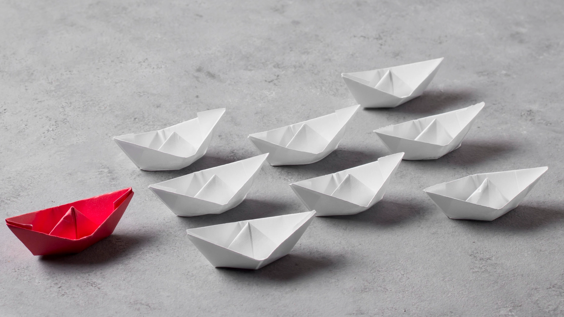 Um grupo de oito barcos de papel de origami em uma superfície cinza, com um barco vermelho liderando o caminho e sete barcos brancos seguindo em formação dispersa. O barco vermelho exemplifica fatores críticos de sucesso, incorporando liderança e singularidade em meio a seus equivalentes. - Efacont