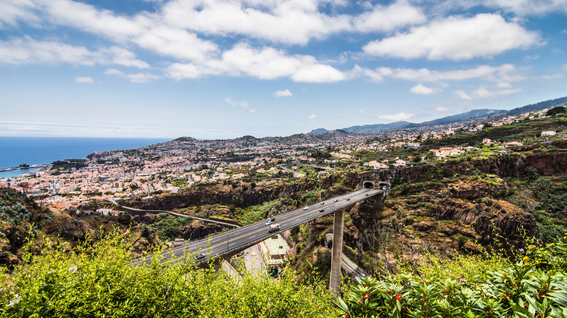 Uma vista aérea de uma paisagem pitoresca em Portugal apresenta uma ponte alta sobre uma ravina profunda, levando a dois túneis. Colinas verdejantes e uma cidade extensa com muitos edifícios a cercam. À distância, montanhas e o oceano azul se encontram sob um céu parcialmente nublado — um verdadeiro indicado ao Destinos e Projetos de Excelência para o World Travel Awards 2024. - Efacont