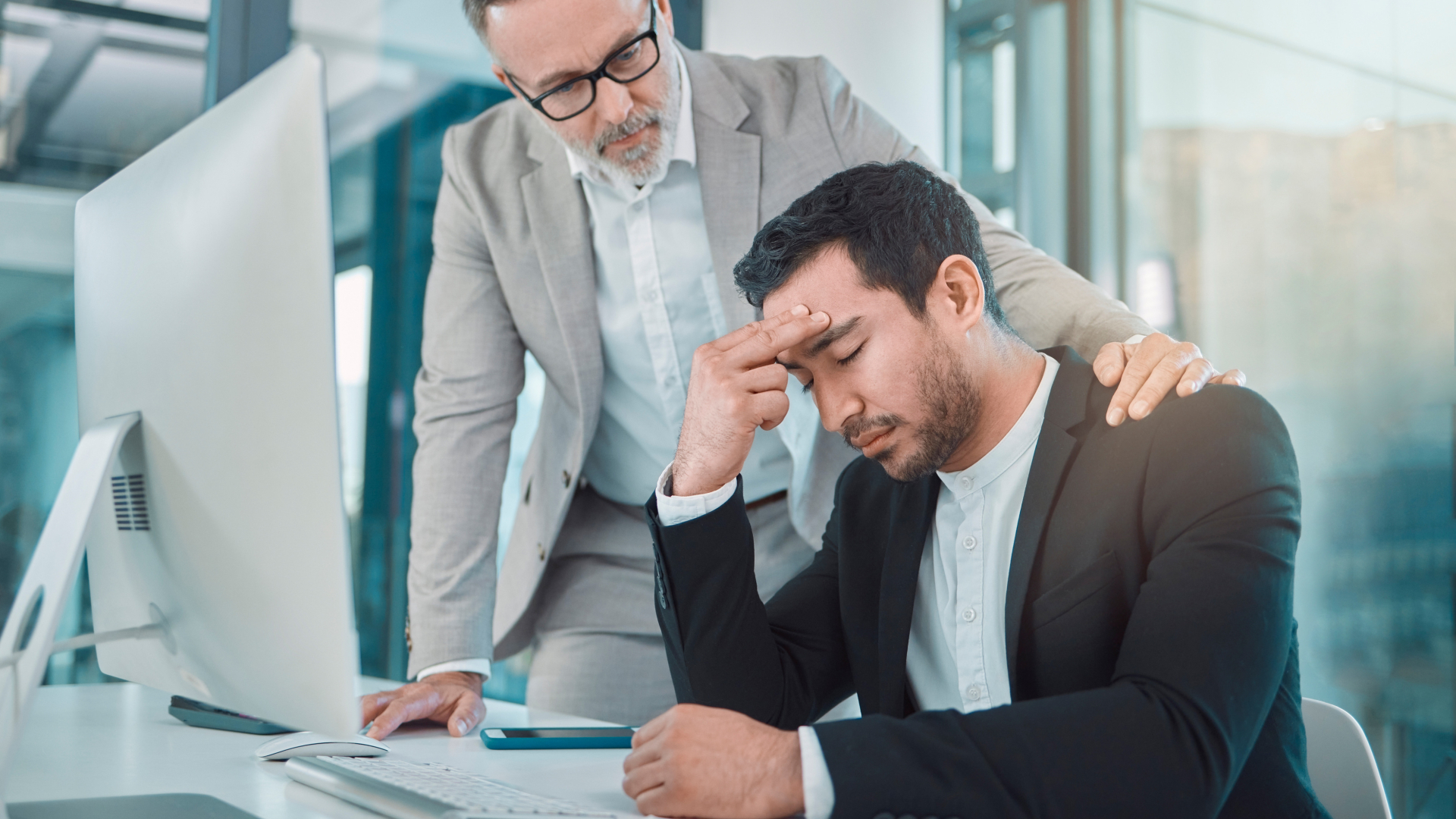 Um homem mais velho em um terno claro se inclina para consolar um homem mais jovem em um terno escuro sentado em uma mesa, aparentemente angustiado pela insolvência iminente. O homem mais jovem descansa a cabeça em uma mão, cercado por um monitor de computador, teclado e smartphone no ambiente de escritório iluminado. - Efacont