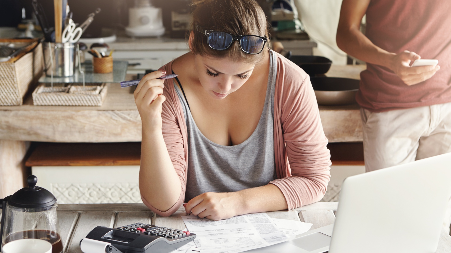 Uma mulher usando óculos na cabeça está sentada à mesa, focada em papéis sobre impostos na sua frente, segurando uma caneta pensativamente. Uma calculadora, cafeteira e laptop aberto estão próximos. Outra pessoa está de pé ao fundo. O cenário parece ser uma cozinha ou um espaço de trabalho casual. - Efacont