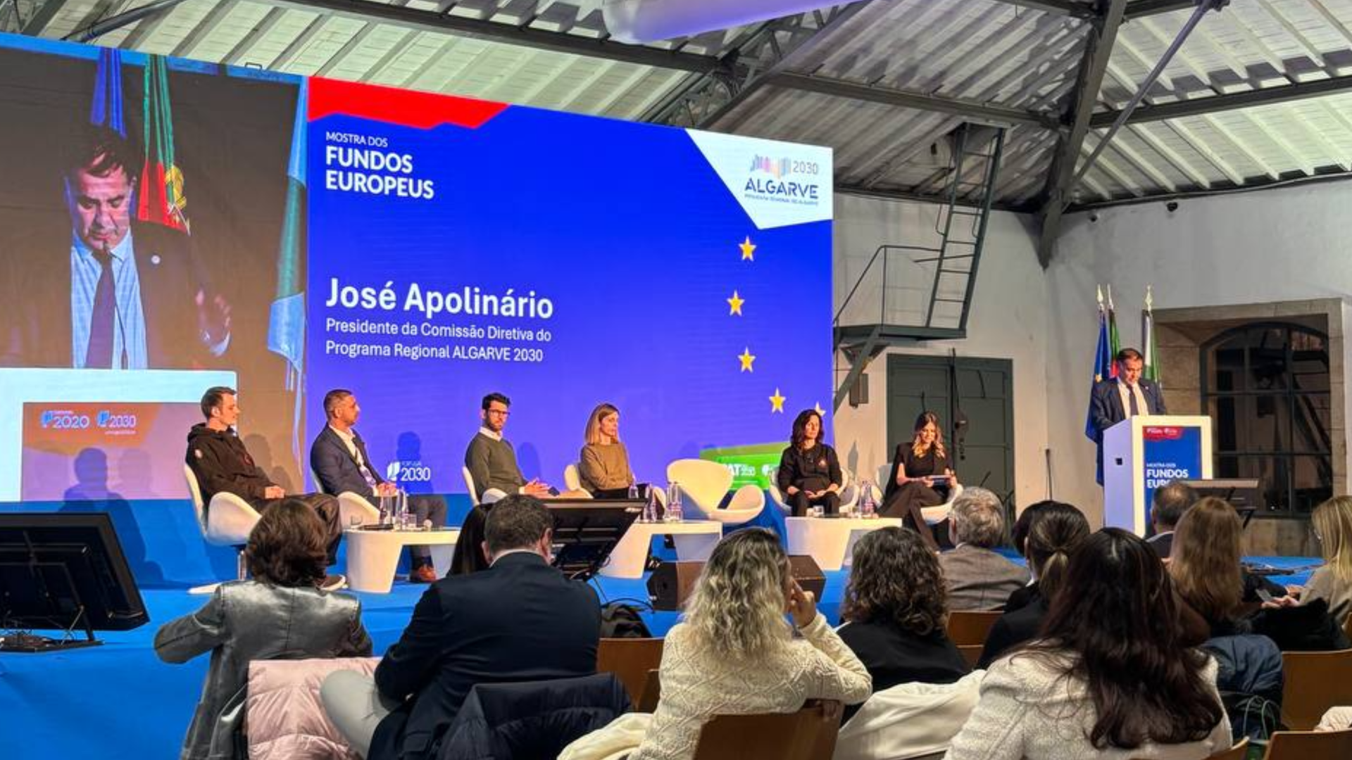 Um painel de discussão na conferência apresenta sete palestrantes no palco sob uma tela grande com "José Apolinário" e detalhes do evento sobre "Mostra dos Fundos Europeus" e "Algarve 2030". O público se senta em fileiras sob um teto alto com vigas expostas. - Efacont