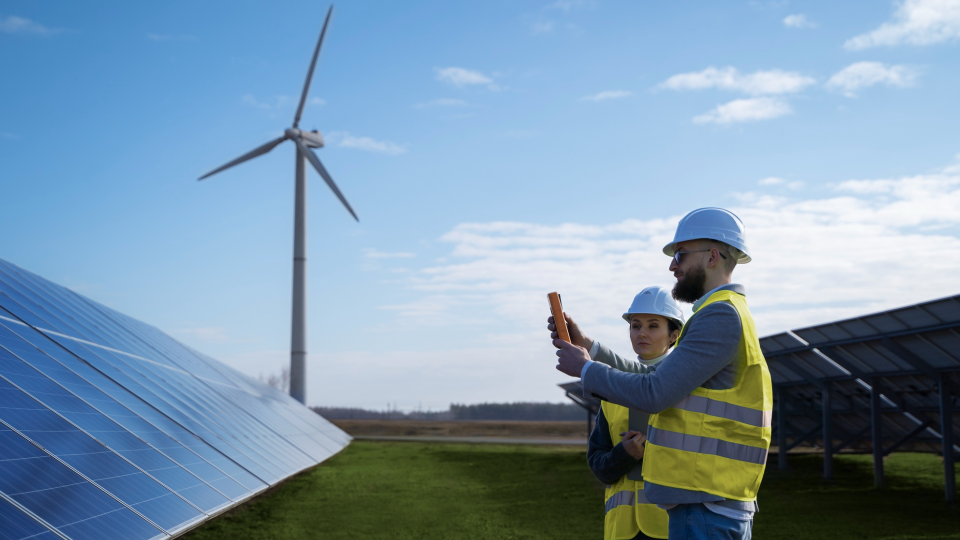 Duas pessoas usando capacetes e coletes refletivos estão em um campo com painéis solares e uma turbina eólica, exemplificando como a produção renovável alcança recorde. Uma pessoa segura um instrumento, possivelmente medindo a produção solar contra o pano de fundo de um céu azul com nuvens dispersas. - Efacont