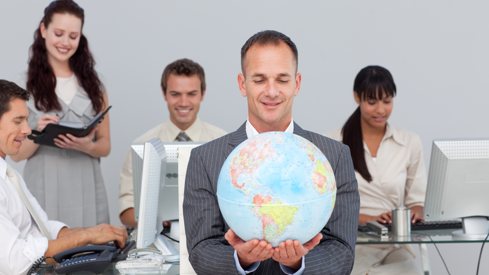 Um homem de terno segura um grande globo enquanto está sentado em uma mesa, cercado por quatro colegas. Dois homens e duas mulheres estão trabalhando em computadores, escolhendo o melhor país para expandir negócio. O escritório moderno exala uma atmosfera colaborativa com todos focados e engajados. - Efacont