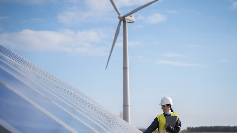 Um trabalhador com capacete e colete de segurança inspeciona painéis solares em primeiro plano, segurando uma prancheta. Uma grande turbina eólica está atrás deles contra um céu azul com nuvens dispersas, simbolizando energia renovável como Produção Renovável Alcança Recorde . - Efacont