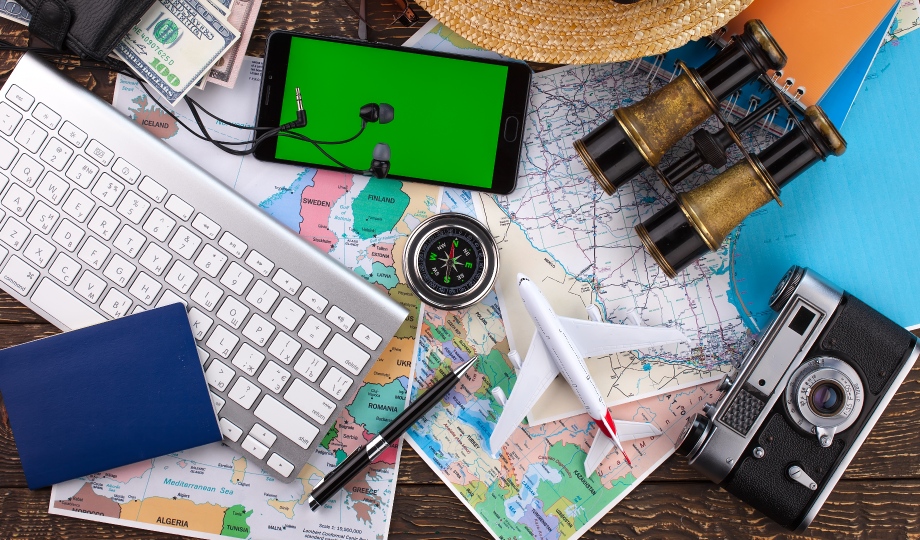 Flatlay com tema de viagem em uma superfície de madeira com mapa, teclado, smartphone com fones de ouvido, binóculos, chapéu de palha, bússola, avião de brinquedo, caneta, passaporte, câmera vintage e notas de dólar americano espalhadas. A cena reflete o crescimento do setor de turismo com sua promessa de planejamento e aventura. - Efacont