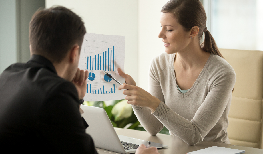 Uma mulher de suéter bege está sentada em uma mesa, apontando para um papel com gráficos de barras e pizza, mergulhando em uma avaliação de empresas. Ela fala com um homem de terno preto, que escuta atentamente. Um laptop está aberto na mesa em meio a um ambiente de escritório com luz natural e plantas verdes ao fundo. - Efacont