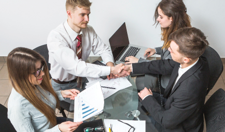 Quatro pessoas em trajes de negócios sentam-se ao redor de uma mesa de vidro, envolvidas em negociações empresariais. Dois homens apertam as mãos, fechando um acordo. Uma mulher com cabelos longos segura um documento exibindo um gráfico de barras, enquanto outra digita em um laptop. Papéis e óculos estão espalhados pela mesa, sinalizando uma reunião de negócios ativa. - Efacont