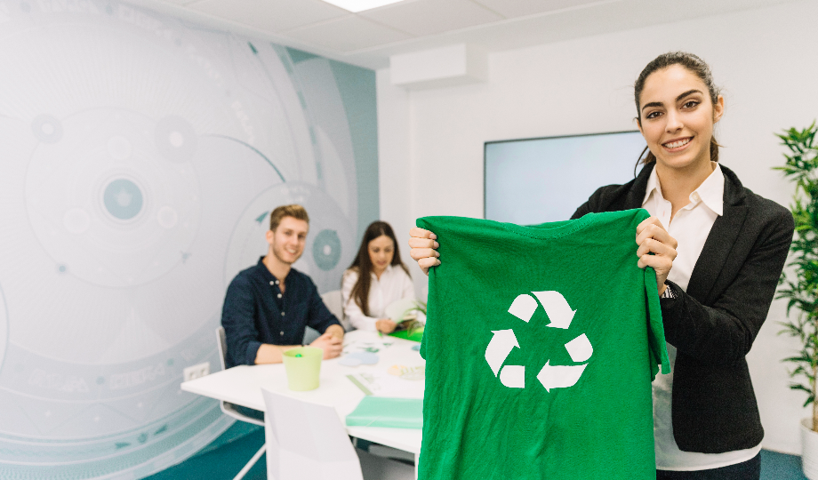 Uma mulher de blazer preto segura uma camisa verde com um símbolo de reciclagem, exibindo sustentabilidade na indústria agrícola. Ao fundo, um homem e uma mulher sentam-se à mesa em um escritório moderno adornado com plantas e decoração com tema de reciclagem, criando uma atmosfera brilhante e ecológica. - Efacont