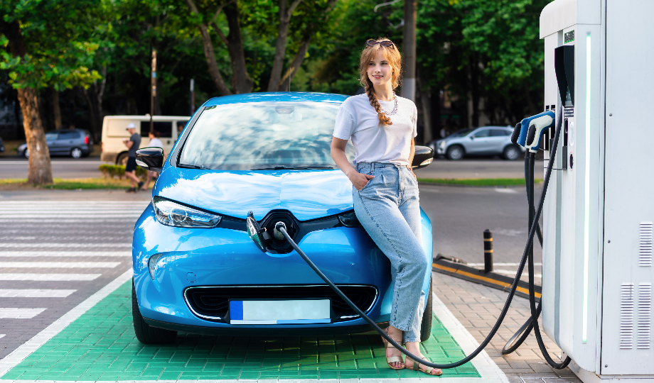 Uma mulher se apoia em um carro elétrico azul em uma estação de recarga, personificando o espírito do incentivo veículos elétricos. Ela veste uma camiseta branca, jeans azul claro e óculos escuros na cabeça, enquanto o cabo carrega seu veículo. No fundo, árvores e tráfego movimentado da rua completam a cena. - Efacont